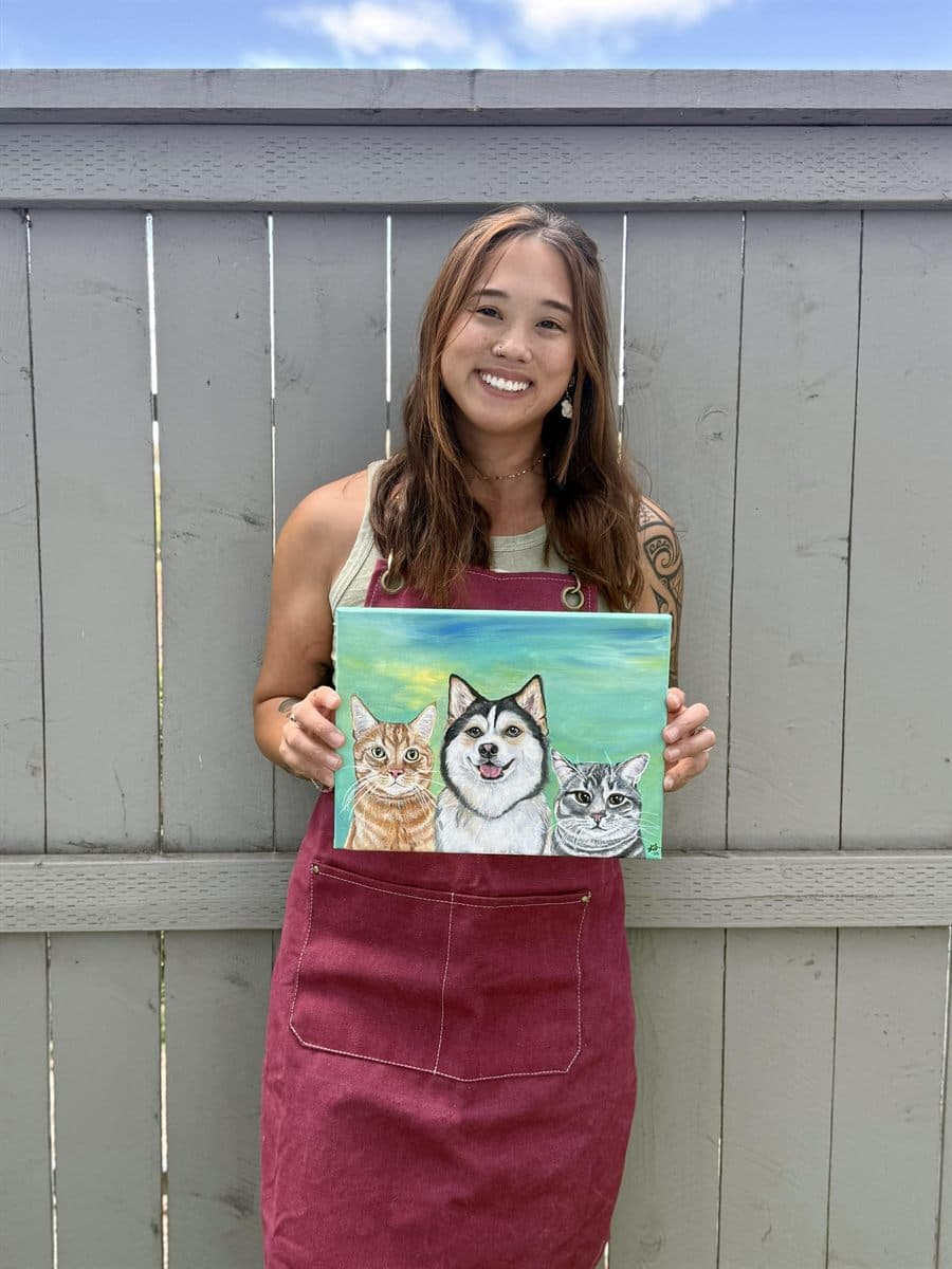 Kim, the artist behind Doodles n Plants, wearing a burgundy apron and holding a hand-painted canvas portrait of three pets, in front of a wood fence