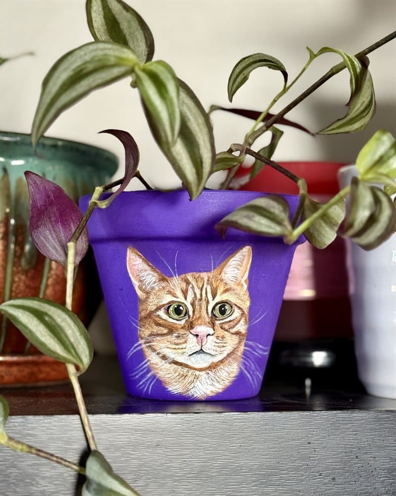 Hand-painted orange tabby cat on a purple clay pot among trailing plants